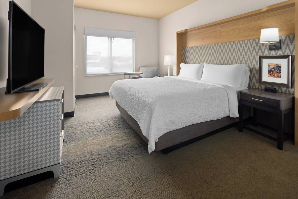 Ein Hotelzimmer mit einem großen Bett, einem Flachbildfernseher, einem Schreibtisch mit Stuhl, einem kleinen Tisch und einem Nachttisch mit Lampe.
