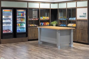 A corner kitchenette with a wooden countertop, a refrigerator stocked with beverages, and shelves with snacks and packaged goods. - Holiday Inn & Suites Savannah Airport - Pooler by IHG (Pooler)
