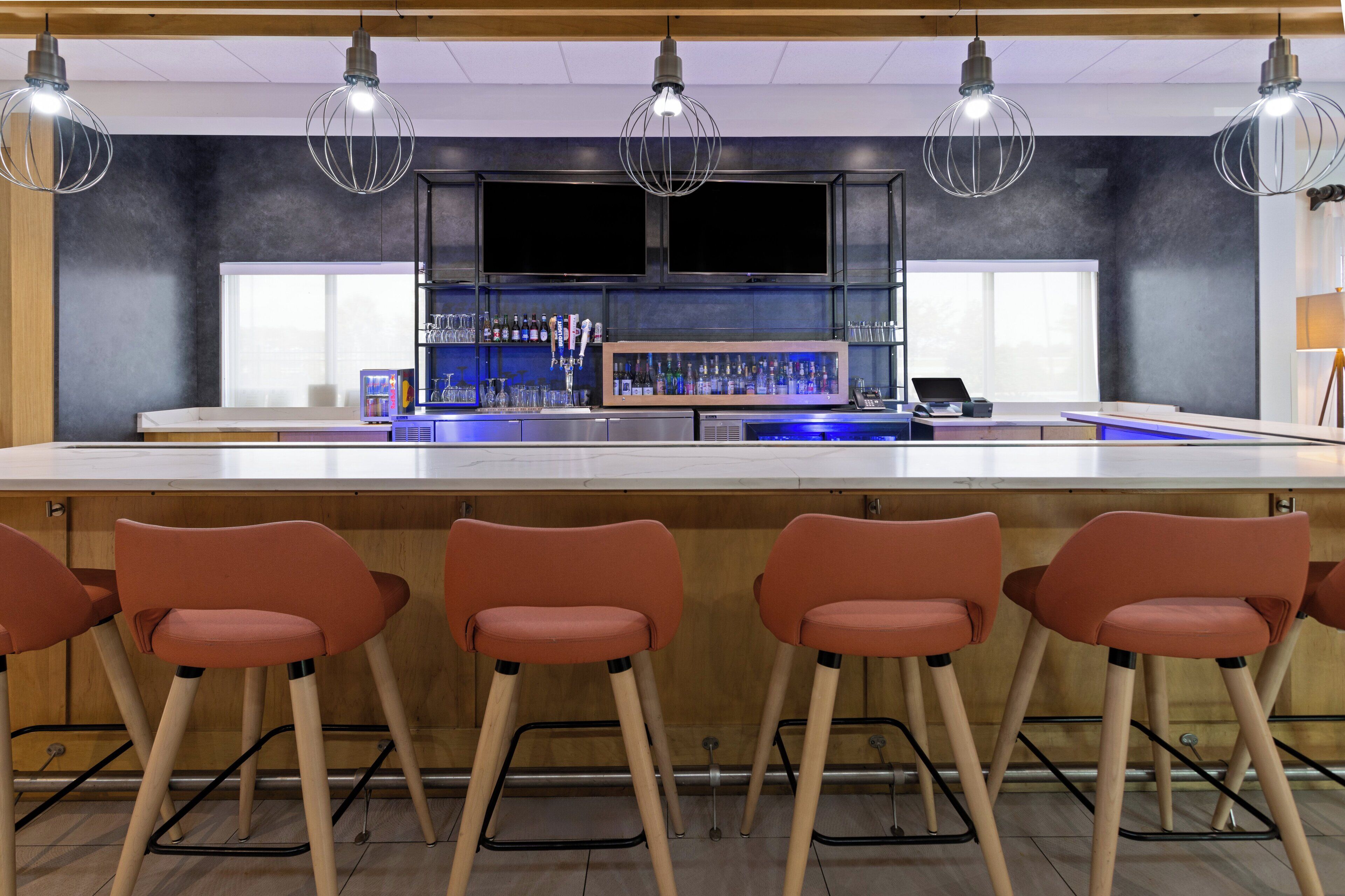 Una zona de bar con cuatro taburetes naranjas, una barra larga y una estantería llena de botellas.