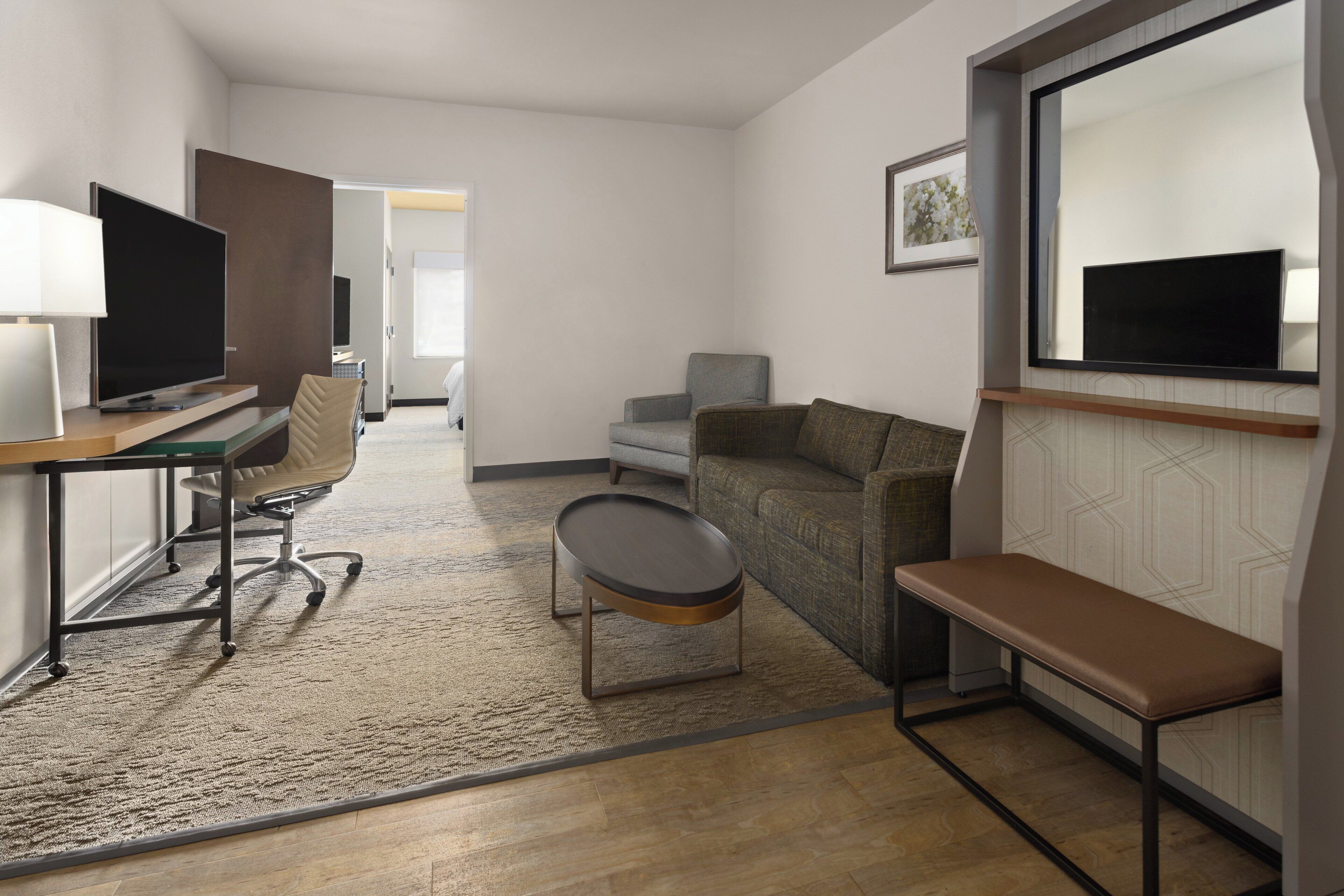 A modern hotel room with a flat-screen TV, a desk, a comfortable sofa, a bench, and a large mirror.
