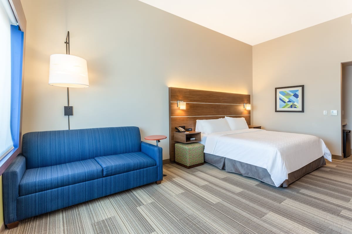 A hotel room with a blue sofa, a bed with white linens, a nightstand with a lamp, and a small table.