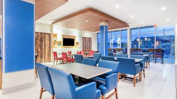 Un comedor moderno con sillas tapizadas en azul y mesas, un televisor de pantalla plana y ventanas amplias con vistas a la ciudad.