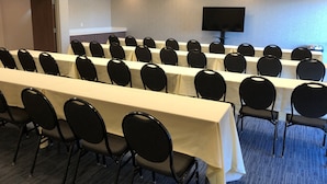 A conference room with rows of tables and chairs, a flat-screen TV on the wall, and a door in the background.