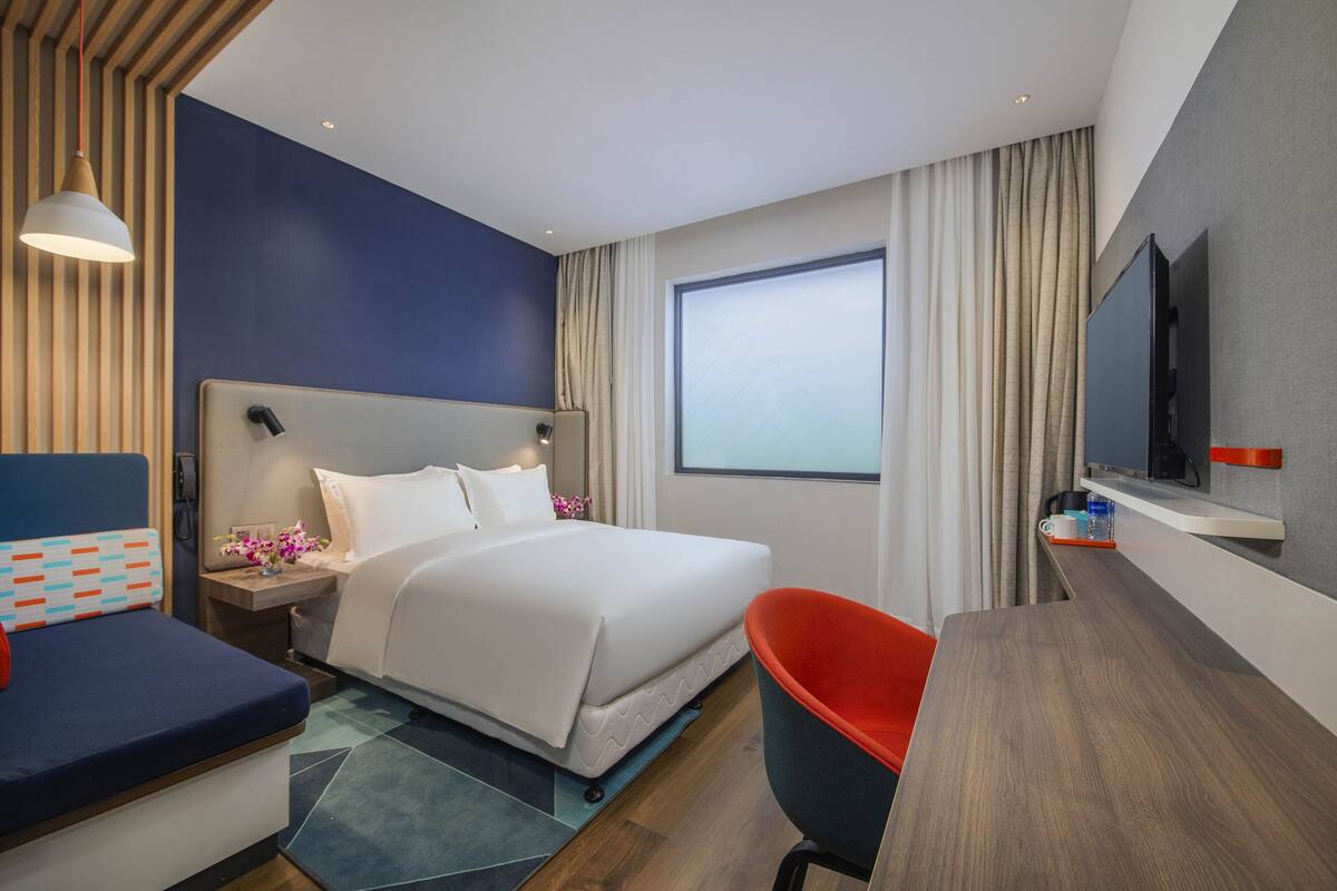 A modern hotel room with a large bed, a desk, a chair, a TV, and a window with curtains.