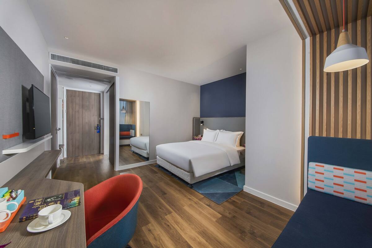 A modern hotel room with a bed, a desk with a chair, a TV, and a window with wooden blinds.
