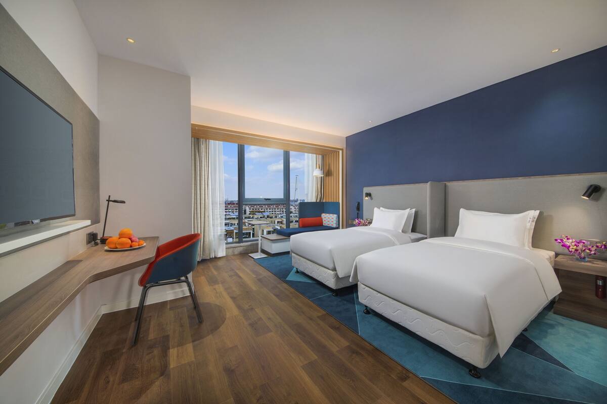 A hotel room with two beds, a desk with a chair, a TV, and a view of the city.