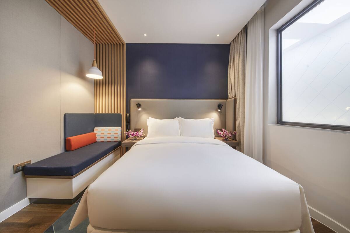 A modern hotel room with a large bed, a built-in bench with cushions, bedside tables with lamps, and a large window offering a view.