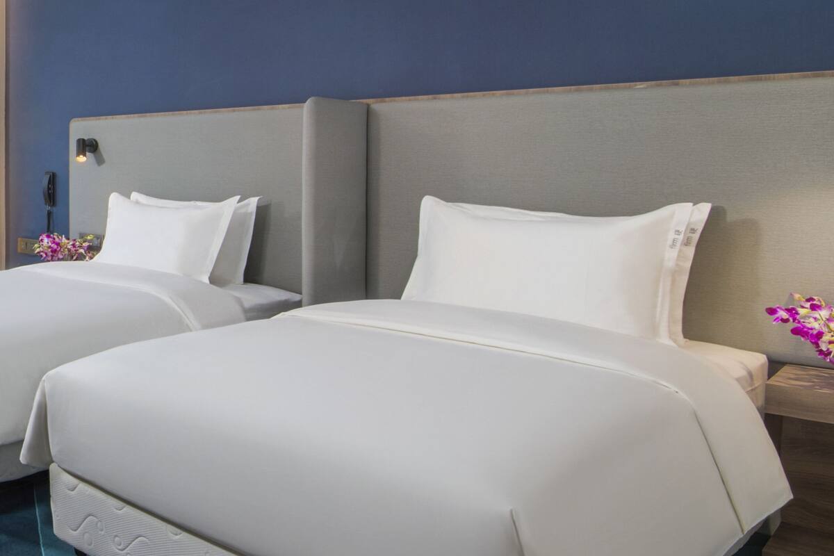 A hotel room with two beds, each with white bedding and pillows. There is a wooden headboard, a small table with a vase of flowers, and a blue wall.