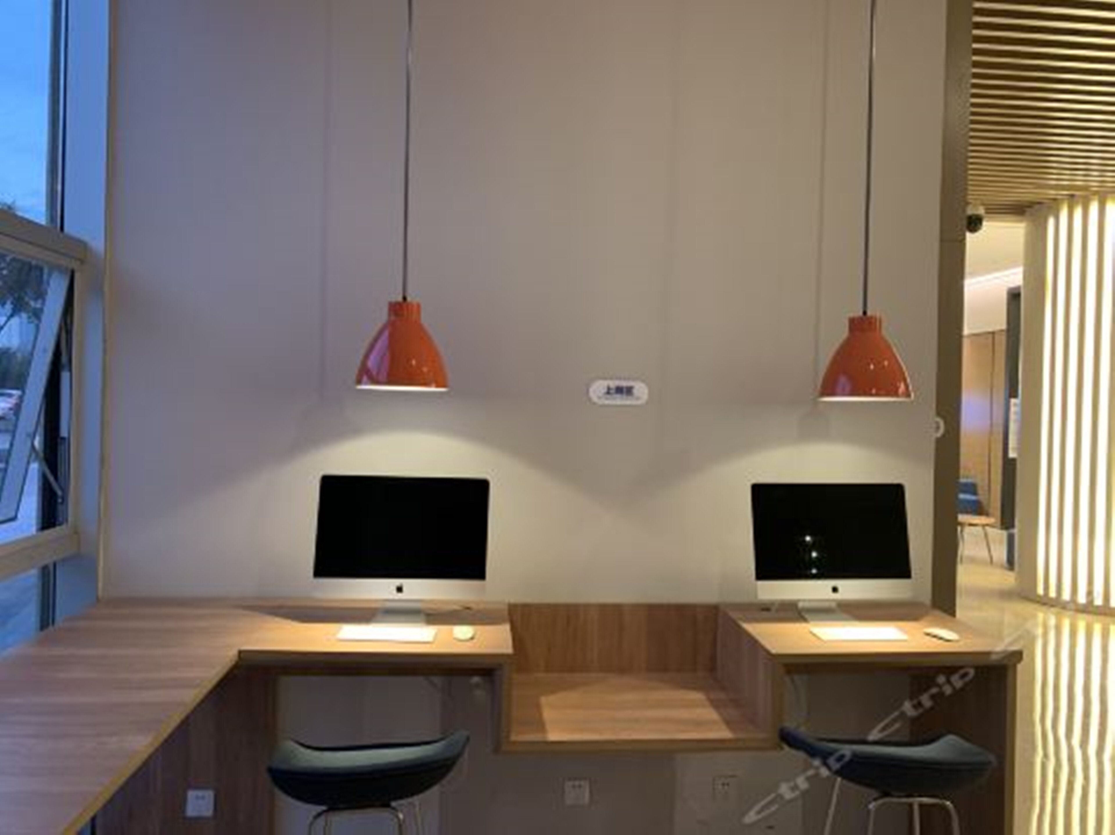 Um espaço de trabalho moderno com duas mesas, cada uma com monitor de computador, teclado e rato. Duas luminárias pendentes laranja estão suspensas acima das mesas.