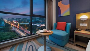 Habitación de hotel con un ventanal grande que ofrece vista a la ciudad al anochecer, un sillón azul con un cojín rojo, una mesita redonda pequeña con una laptop y una taza, y una lámpara de pared.
