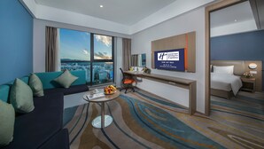 Habitación de hotel con un ventanal amplio que ofrece vistas a la ciudad, un sofá con cojines color turquesa, un escritorio con una silla, una cama y un televisor empotrado en la pared mostrando información del hotel.