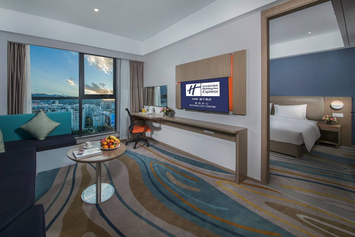 A hotel room with a large window offering a city view, a sofa with teal cushions, a desk with a chair, a bed, and a wall-mounted TV displaying hotel information.