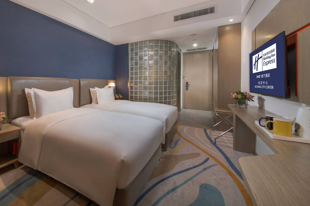 A hotel room with two beds, a desk, a chair, a TV, and a decorative wall.