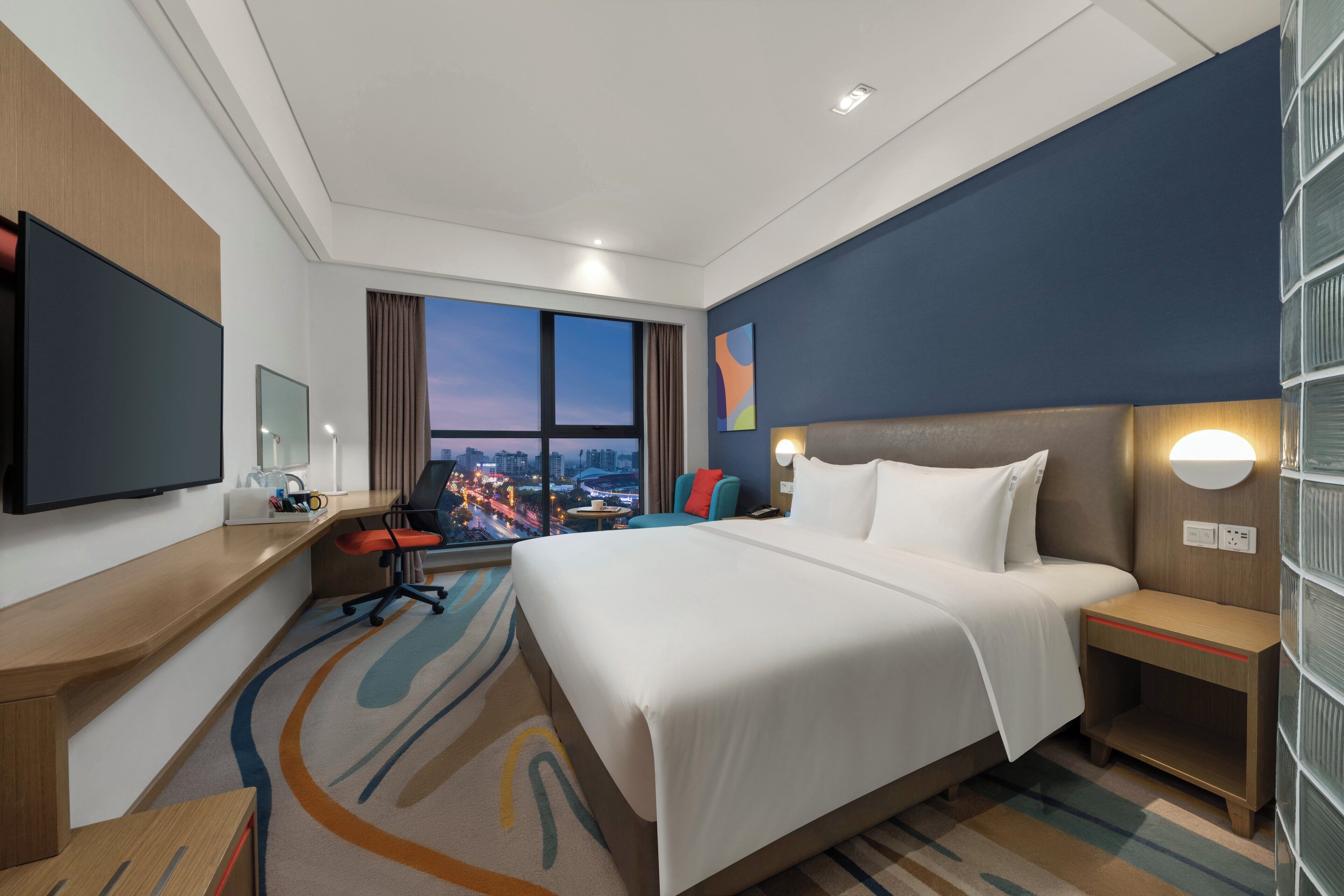 A hotel room with a large bed, a desk, a television, and a view of the cityscape.
