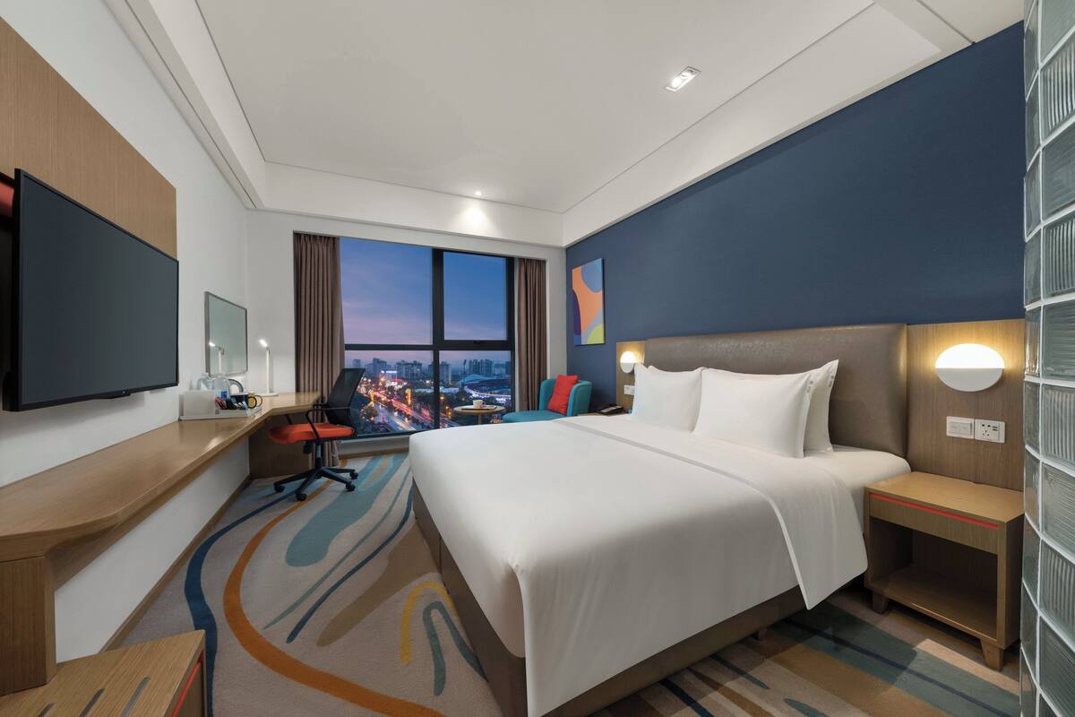 A hotel room with a large bed, a desk, a television, and a view of the cityscape.