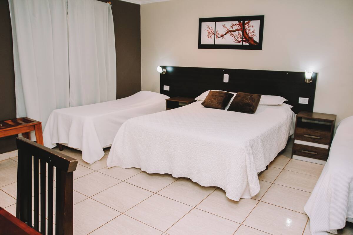 A hotel room with two beds, a wooden chair, a nightstand, and a wall-mounted lamp.