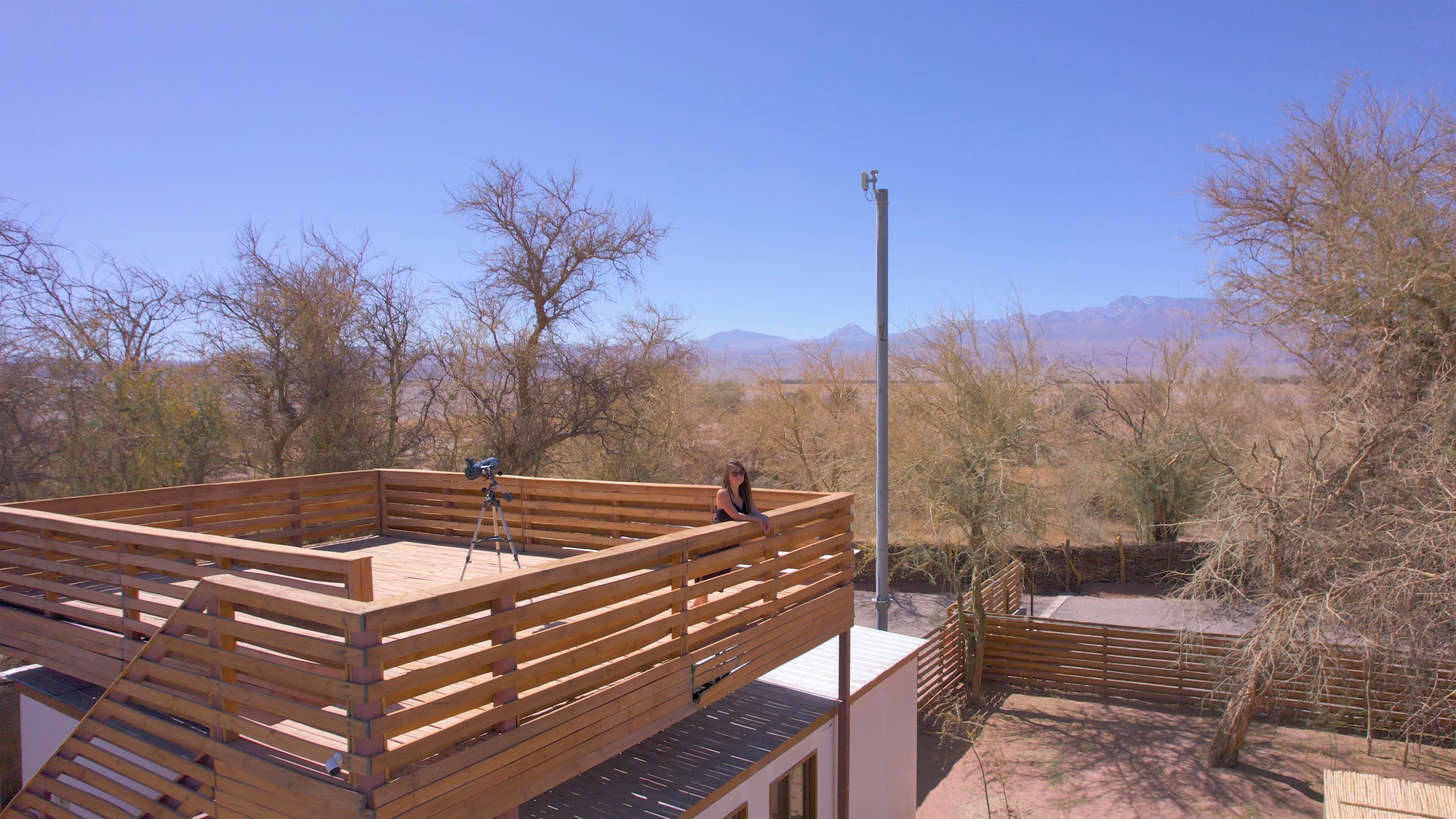 Una terraza de madera con un telescopio, rodeada de árboles y montañas al fondo.