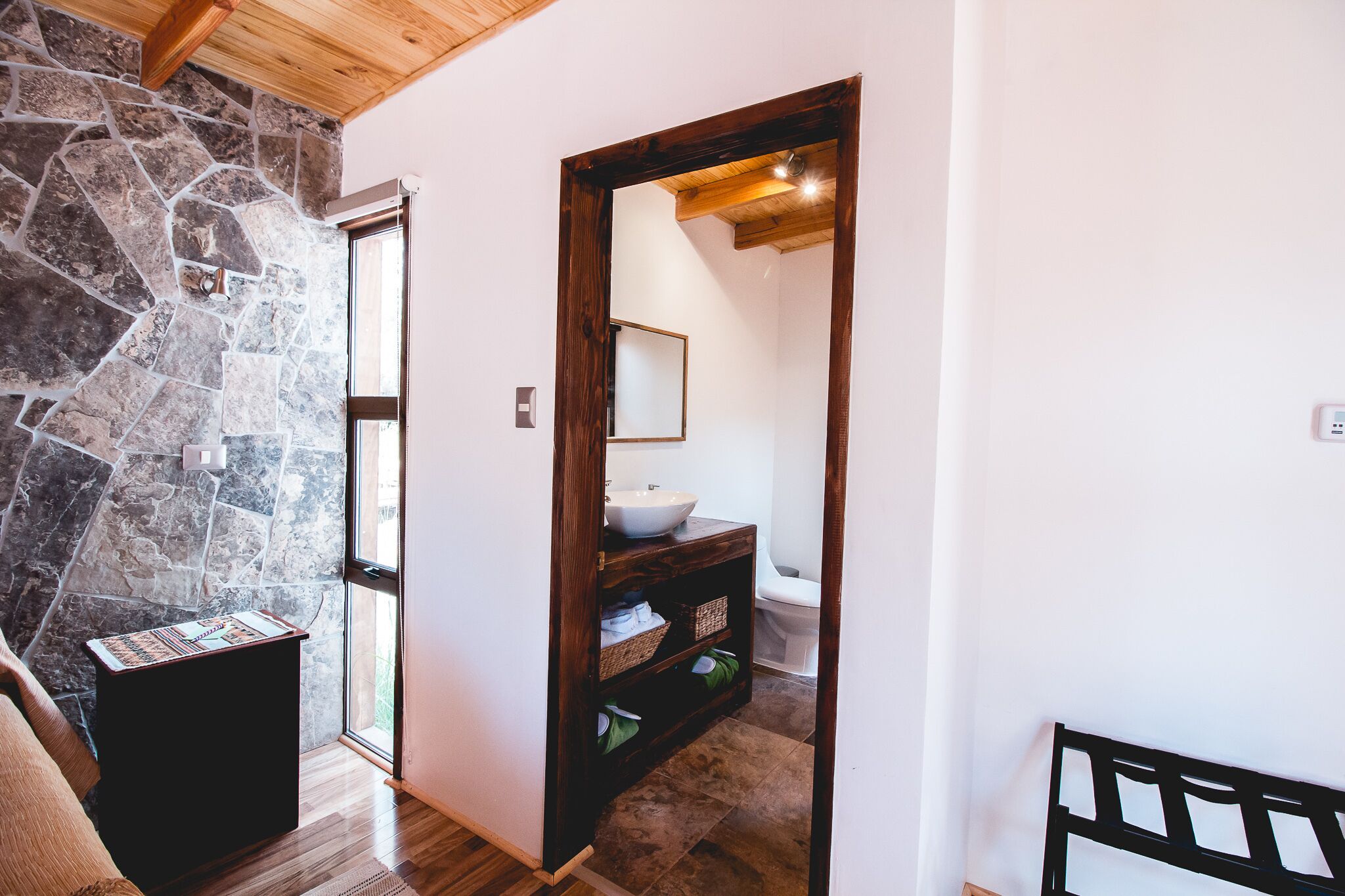 Un couloir avec un mur en pierre, un banc en bois et une porte en bois donnant sur une salle de bain équipée d’un lavabo et d’un miroir.