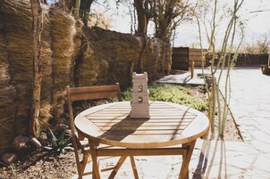Una mesa de madera redonda con un farol de piedra en el centro, rodeada de sillas, ubicada al aire libre con un muro de piedra y vegetación de fondo.