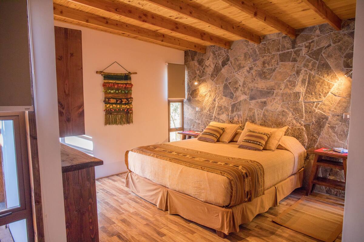 Une chambre avec un mur en pierre, des poutres en bois au plafond, un lit recouvert de plusieurs coussins, une petite table et une tapisserie murale.