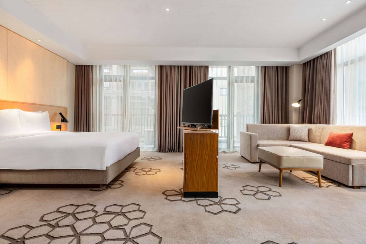 Una habitación de hotel moderna con una cama grande, un sofá, un televisor de pantalla plana sobre un atril de madera y una alfombra estampada.