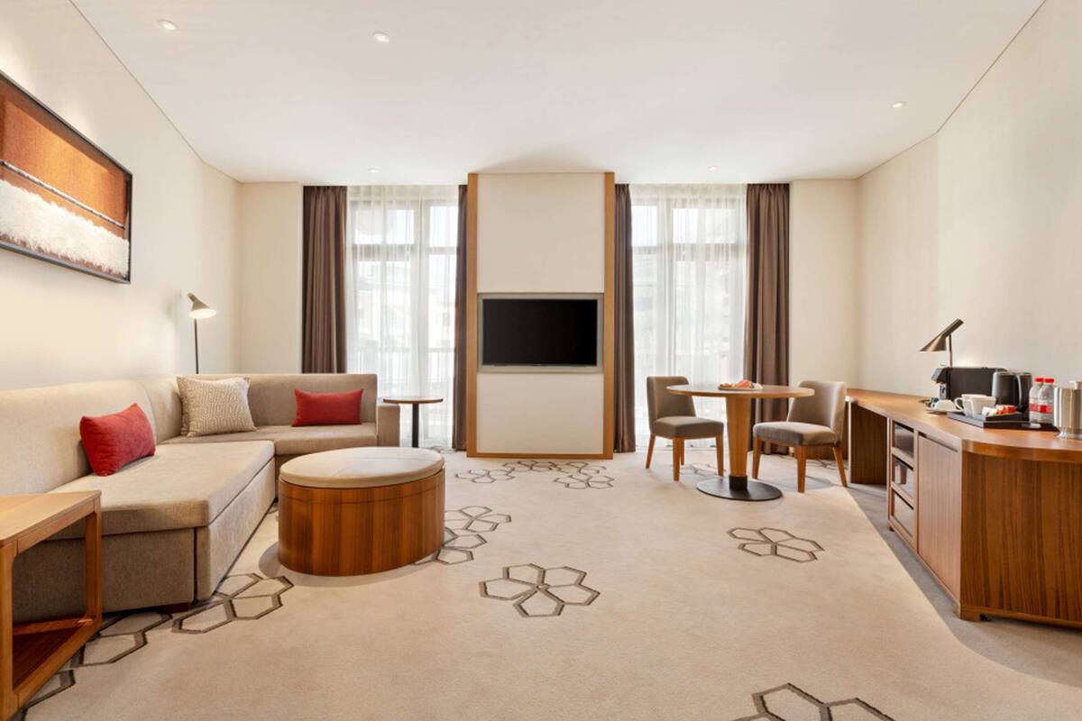 Una habitación de hotel moderna con sofá, una mesita redonda, televisión, escritorio con silla y zona de cocina.