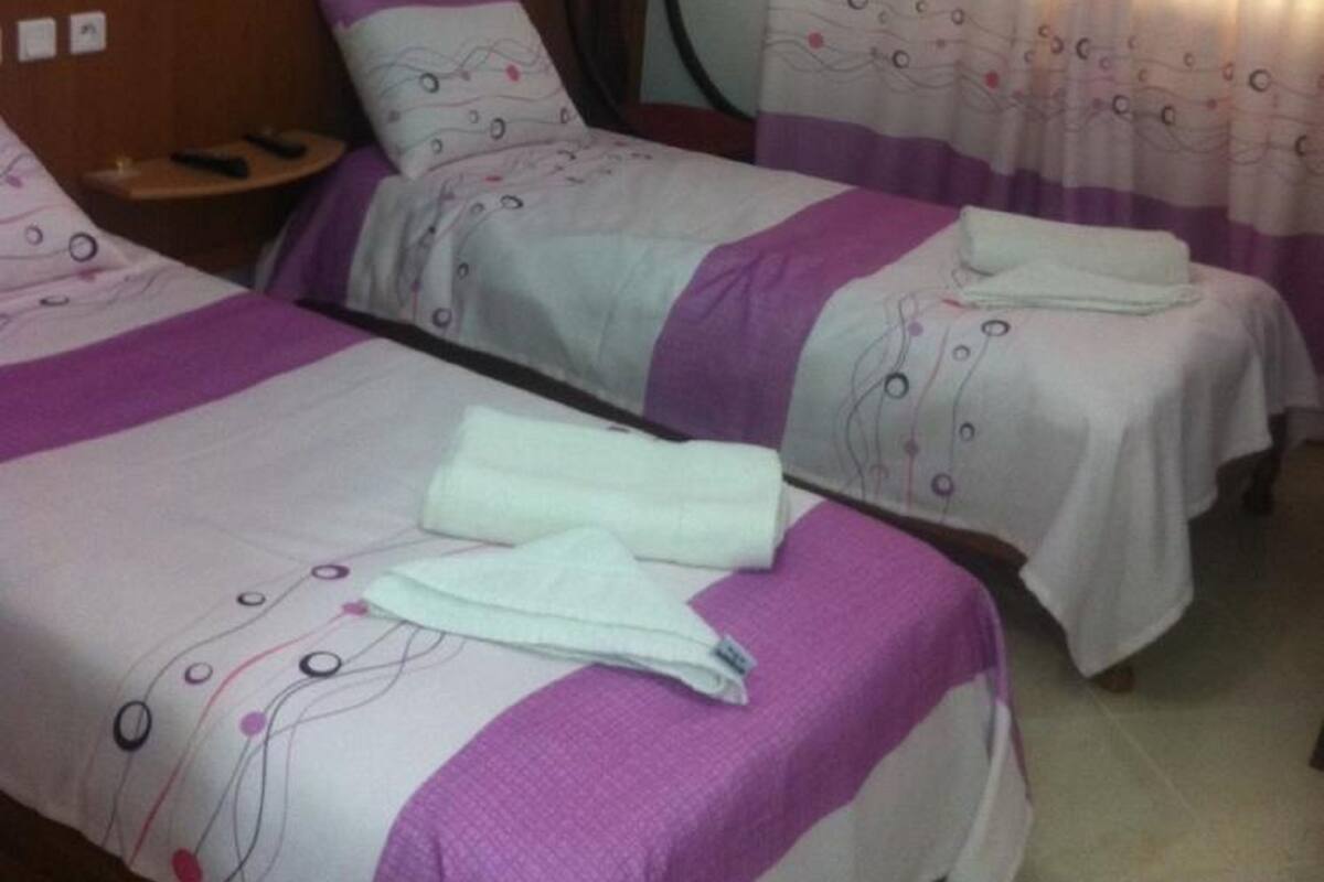 A hotel room with two beds, each with a headboard and a pillow. There is a small table with a remote control on it.