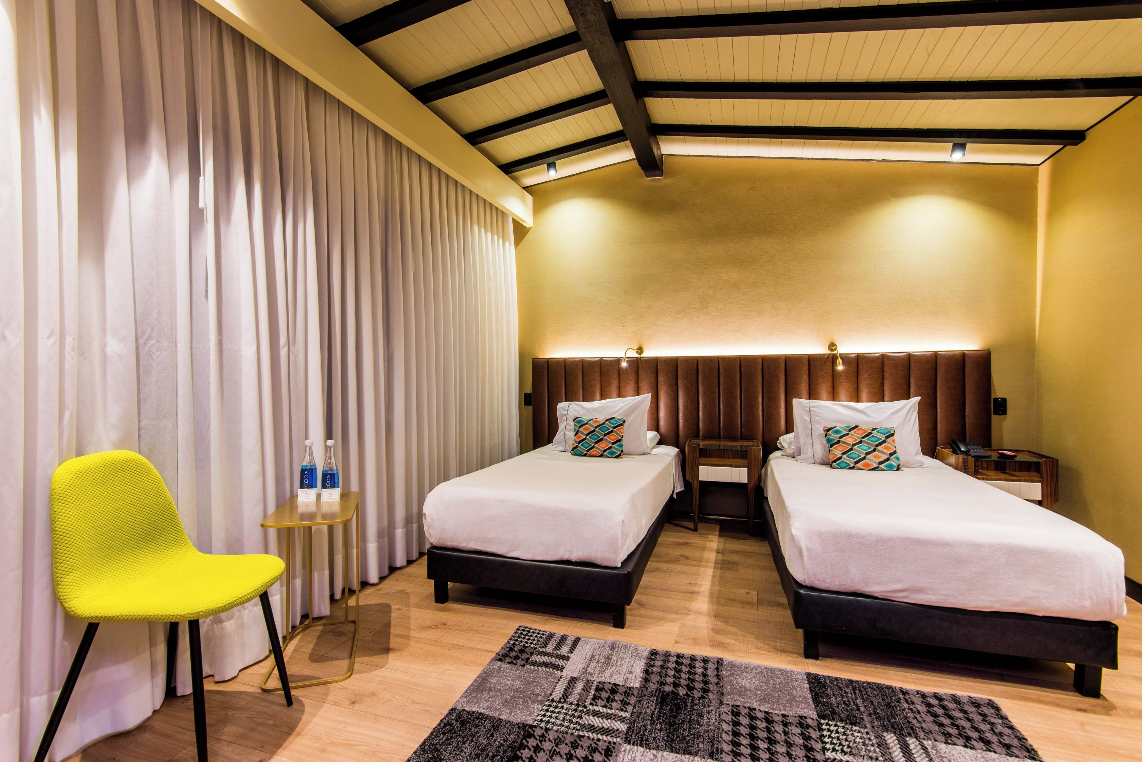 A hotel room with two beds, a yellow chair, a small table with bottles, and a patterned rug.