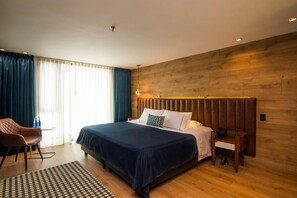 A modern hotel room with a large bed, a wooden headboard, a desk with a chair, and a patterned rug.