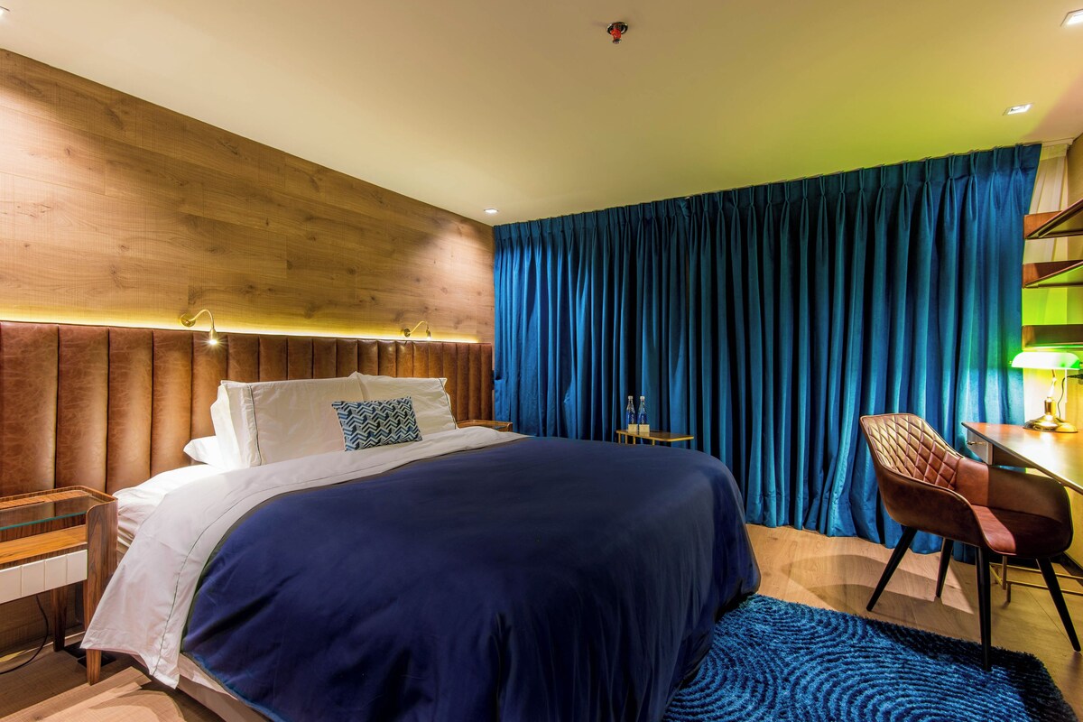 A modern hotel room with a large bed, a wooden headboard, a desk with a chair, and a blue curtain.