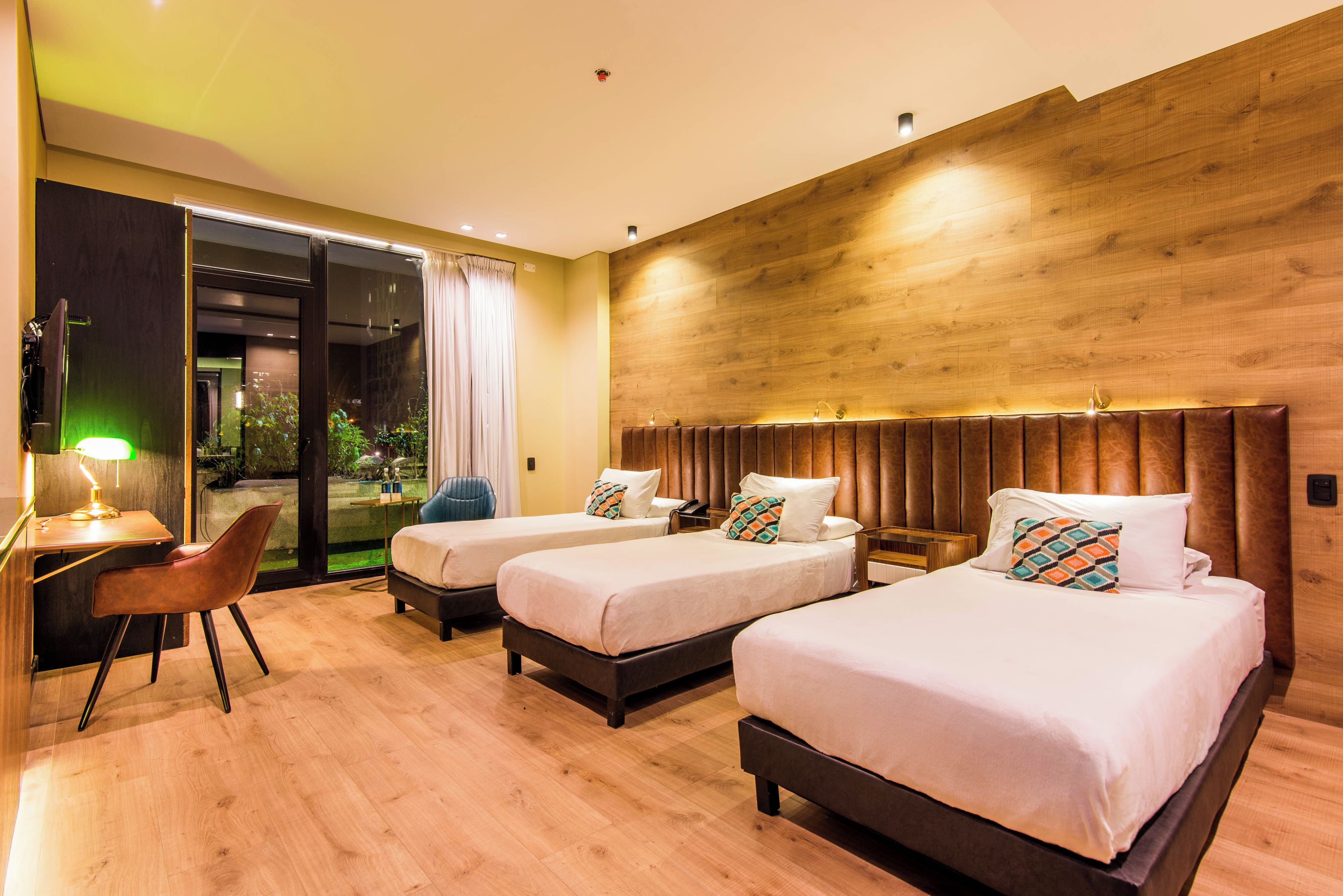 A hotel room with three beds, a desk with a chair, and a large window with curtains.