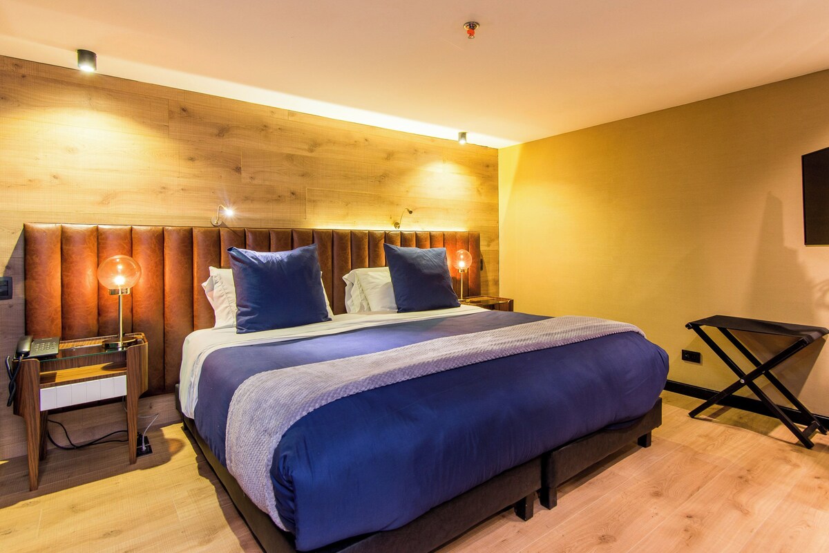A modern bedroom with a wooden headboard, a large bed with blue bedding, bedside lamps, and a foldable table.