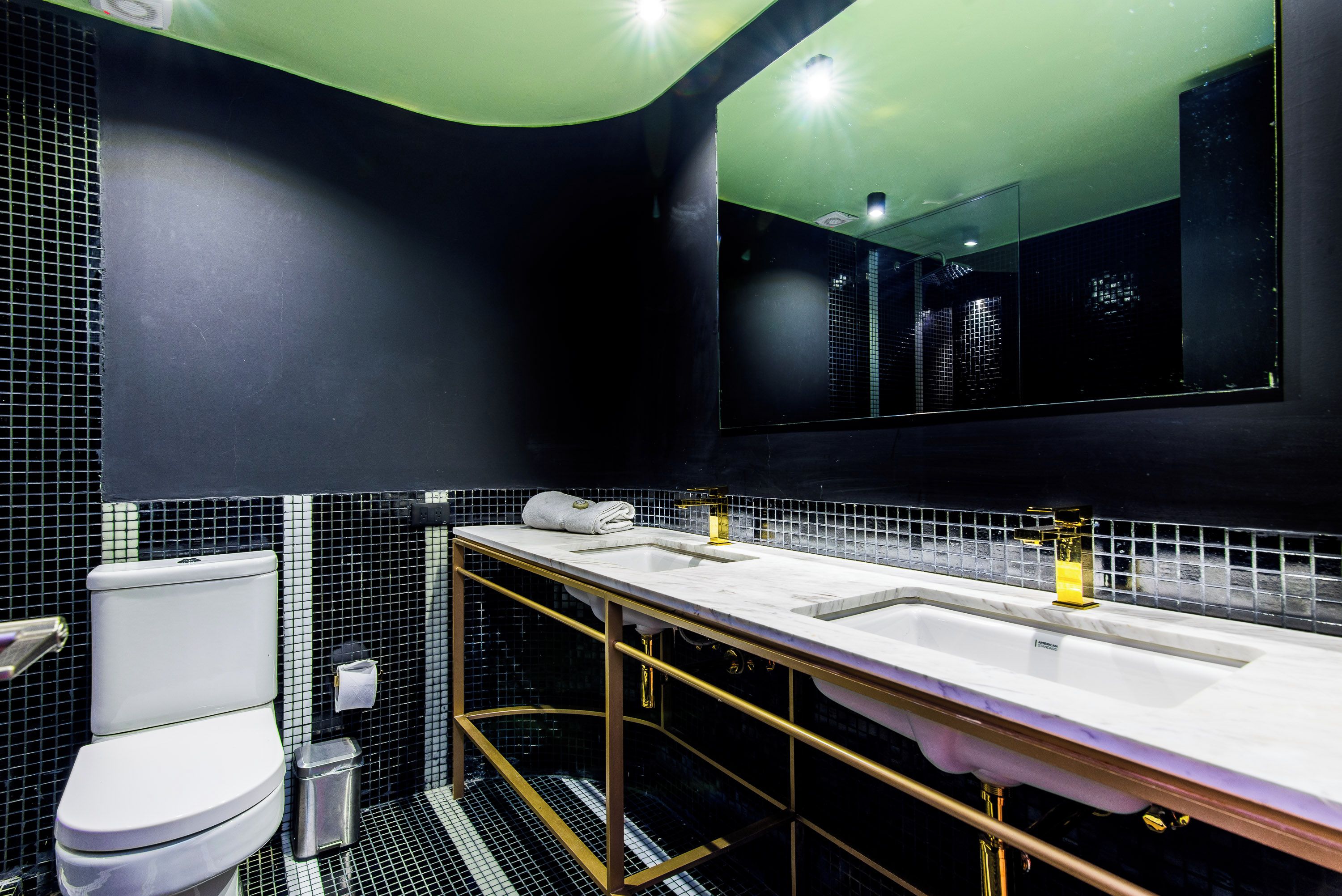 A modern bathroom with a black and white tiled backsplash, a white toilet, a trash can, and a double sink with gold fixtures.