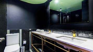 A modern bathroom with a black and white tiled backsplash, a white toilet, a trash can, and a double sink with gold fixtures.