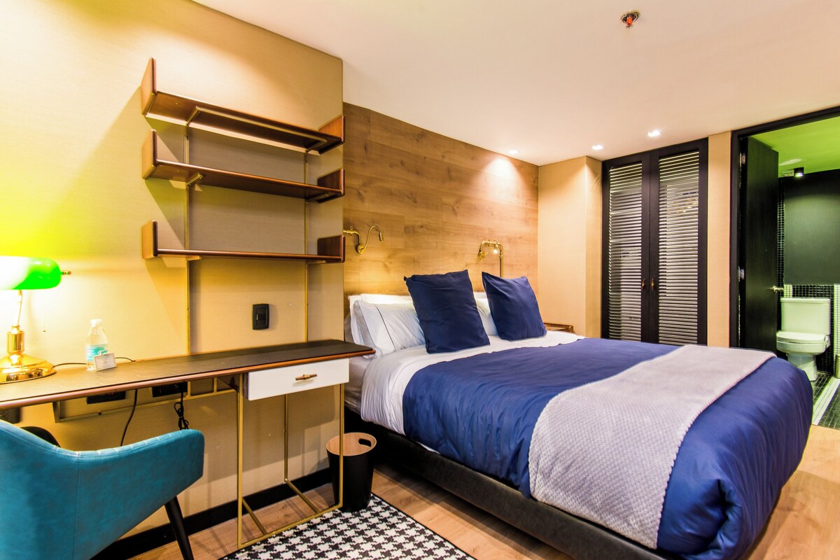 A modern hotel room with a bed, desk, chair, and a bathroom visible through a glass door.