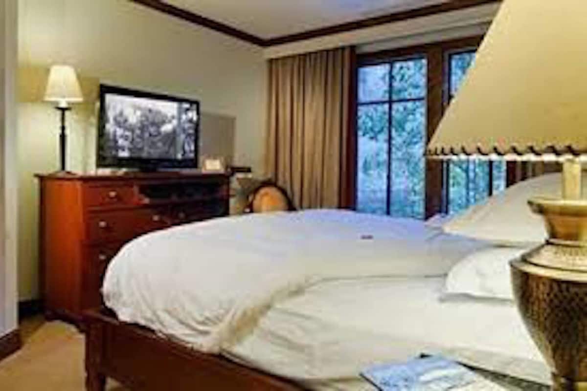 Quarto de hotel com cama, televisão, luminária e janela com cortinas.