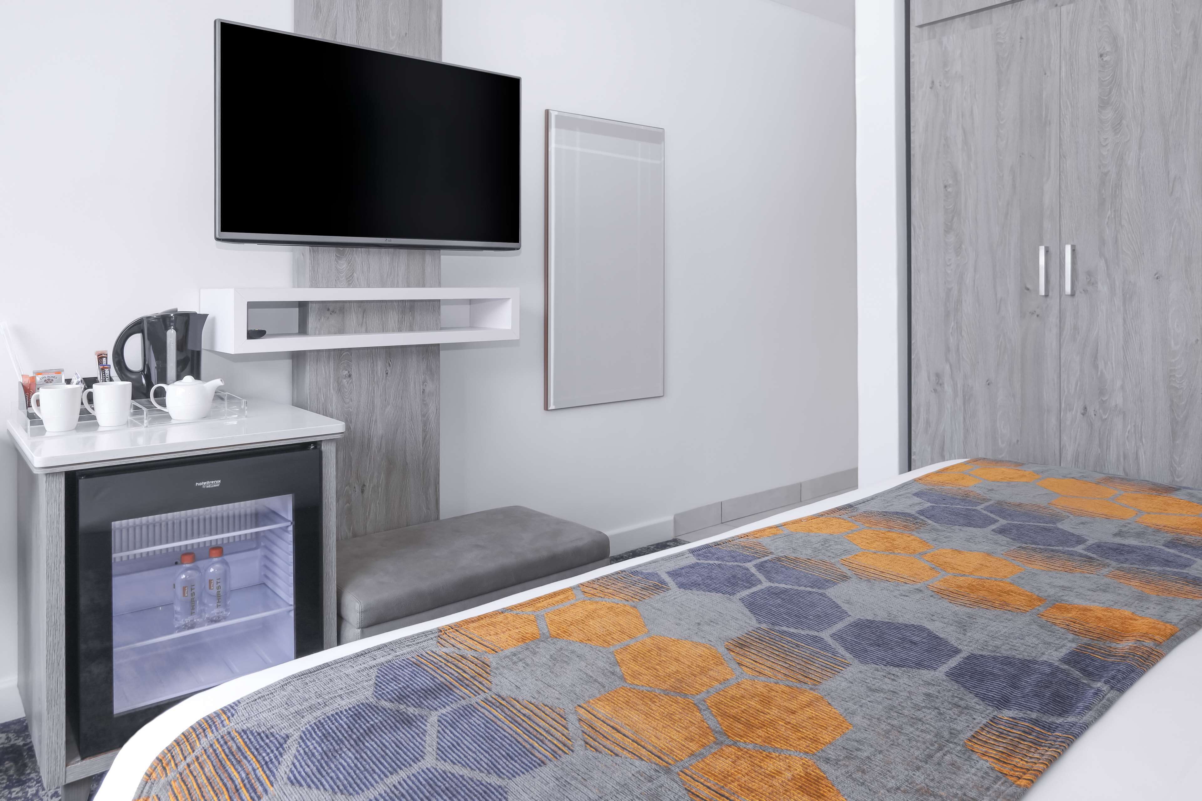 A hotel room with a bed, a TV mounted on the wall, a small fridge, a desk with a kettle, and a wardrobe.