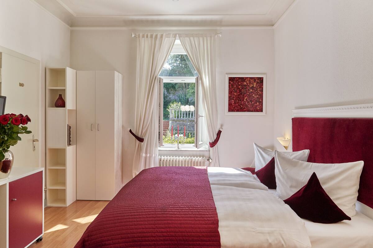 A bedroom with a bed, white curtains, a window with a view, a red vase, and a red headboard.