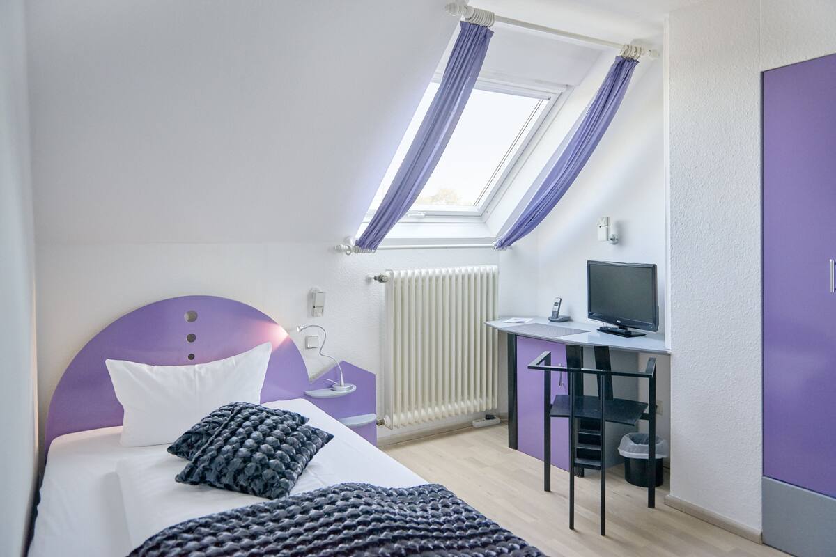 A bedroom with a bed, a desk, a television, and a window with curtains.