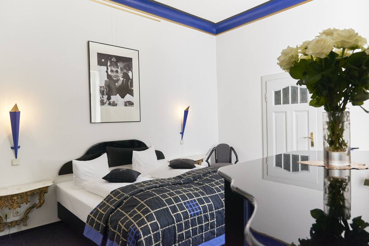 A hotel room with a neatly made bed, a framed photo on the wall, a vase with flowers, and a door with a window.