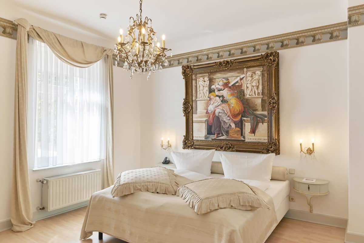 A bedroom with a large bed, a chandelier, a painting, and a nightstand.
