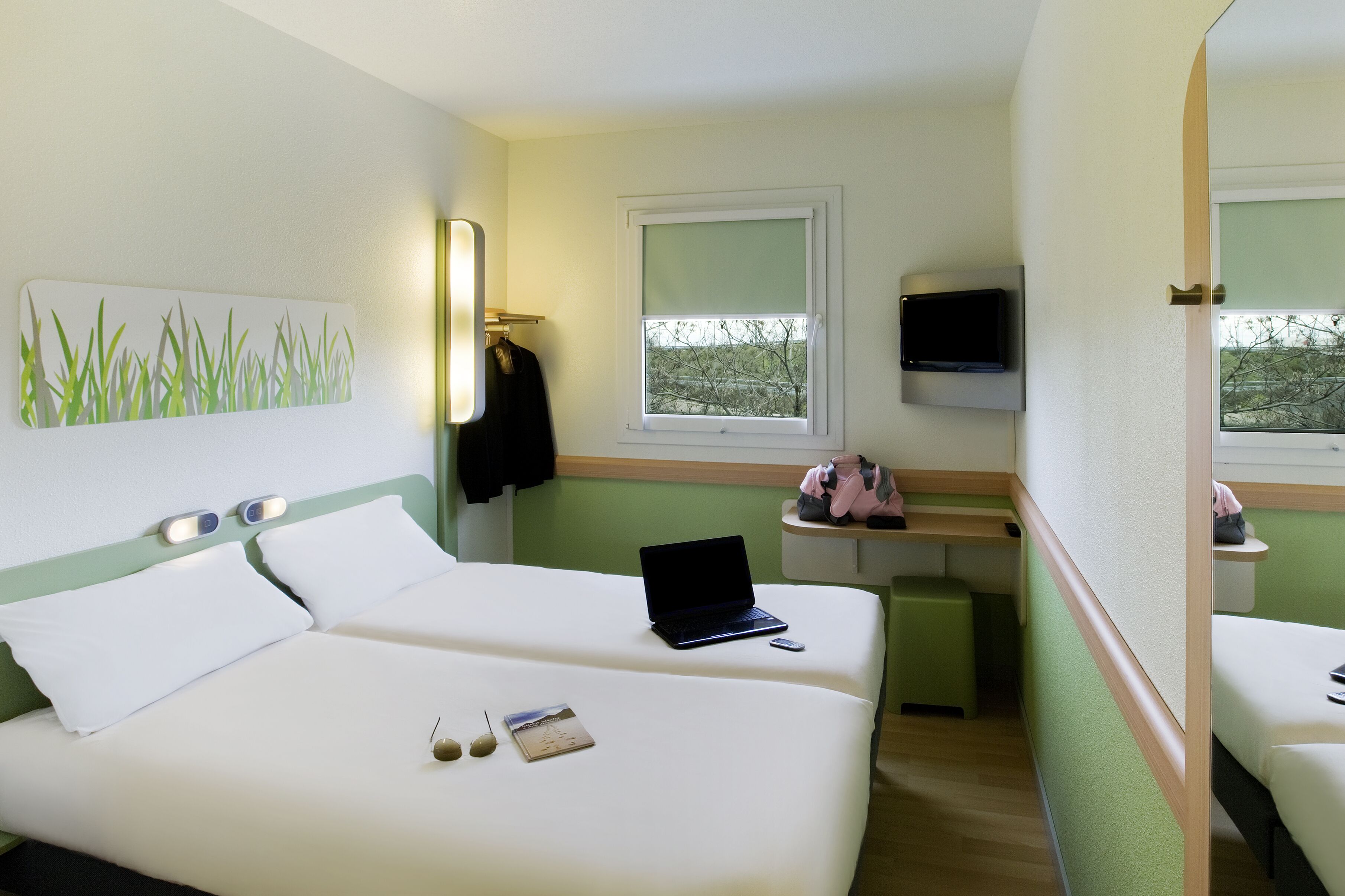 A hotel room with two beds, a laptop on one bed, a TV on the wall, and a window with a view of trees.