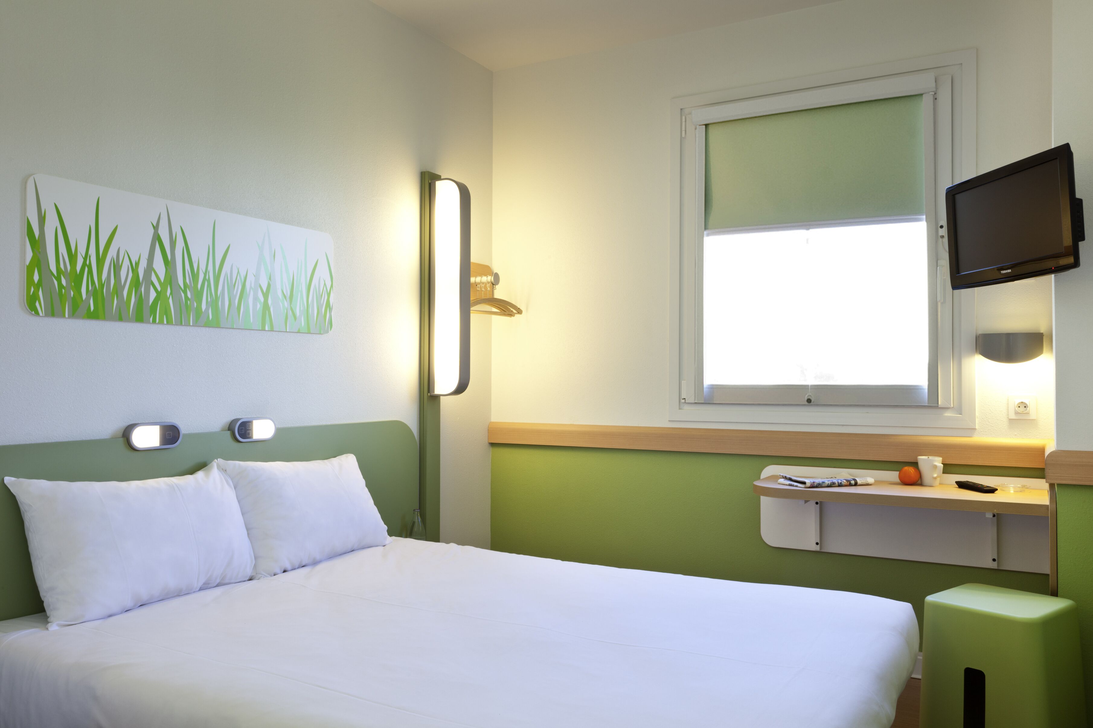 Ein Hotelzimmer mit Bett, Wand-Fernseher, Fenster mit Jalousien und Nachttisch mit Tasse und Fernbedienung.