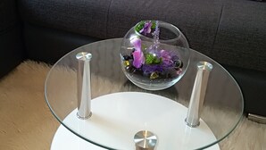 Ein Tisch mit Glasplatte und weißem Fuß, eine Couch mit türkisfarbenen und violetten Kissen und ein flauschiger beiger Teppich.
