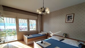 Ein Schlafzimmer mit einem großen Bett, einem kleineren Bett und einer Bank. Ein Fenster bietet Blick auf Grünflächen und es gibt einen Balkon.