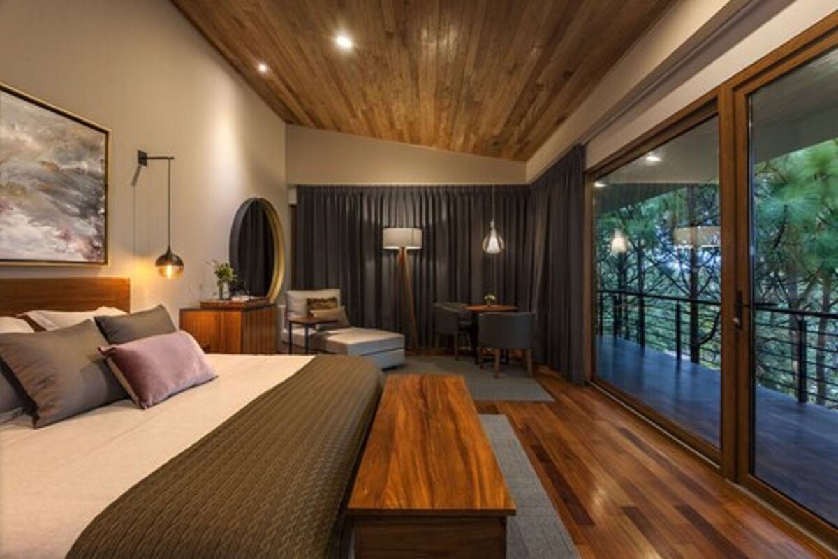 A modern bedroom with a large bed, a wooden bench, a desk, and a view of the outdoors through large windows.