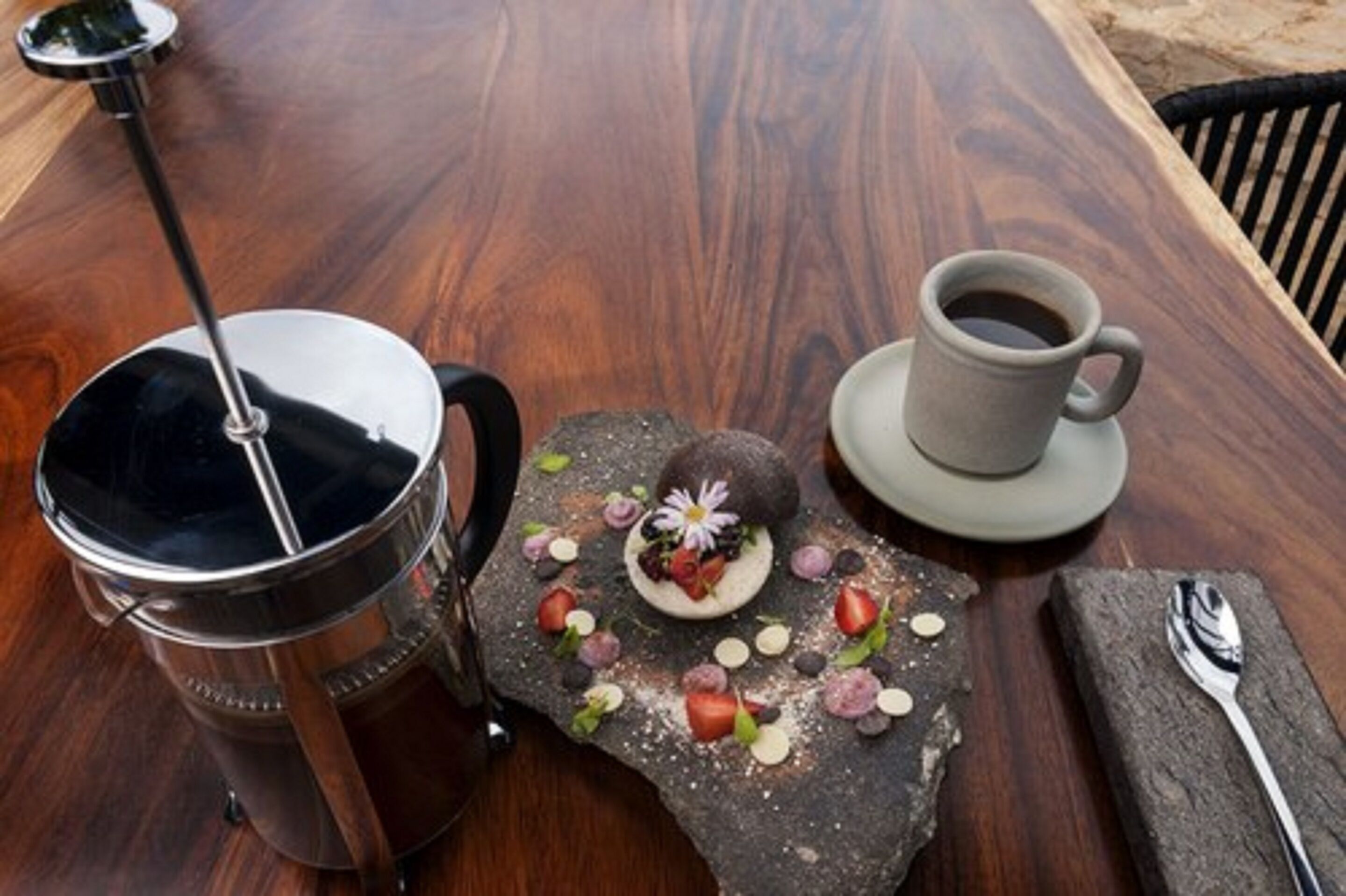 Taula de fusta amb una cafetera, una tassa de cafè i un postre amb maduixes i un flòwer sobre un plat de pedra.