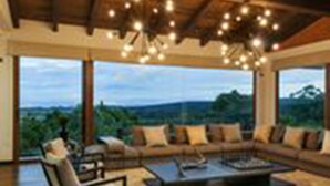 A spacious living room with a large sofa, a coffee table, and a fireplace. There are large windows offering a view of the outdoors.