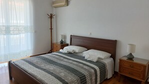Ein ordentlich bezogenes Bett mit gestreifter Bettdecke, zwei Nachttische mit Lampen, ein Holzboden und ein Fenster mit durchscheinenden Vorhängen.