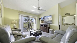 A living room with leather sofas, a ceiling fan, a TV, and a view of palm trees through the sliding glass door.
